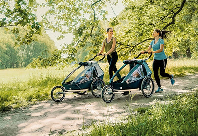 Two women jogging with Croozer Kid Vaaya and Keeke child bike trailers in a park setting