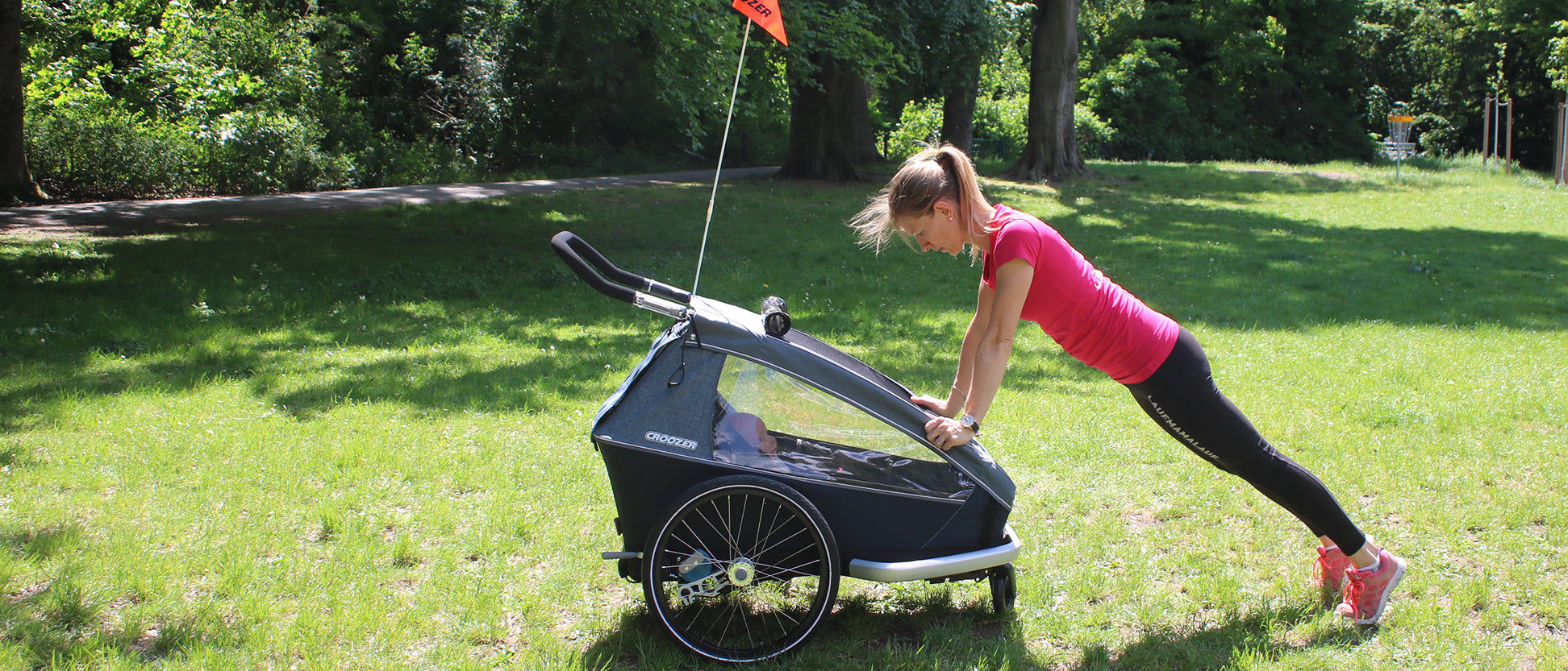Woman in plank position gripping Croozer bike trailer front, exercising outdoors. 