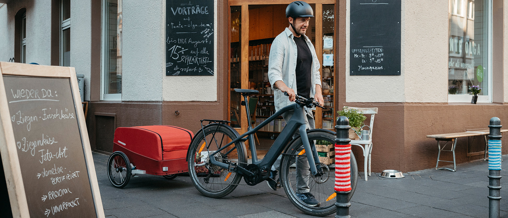 Man pushes bike with Croozer Cargo trailer past a traditional small grocery store.