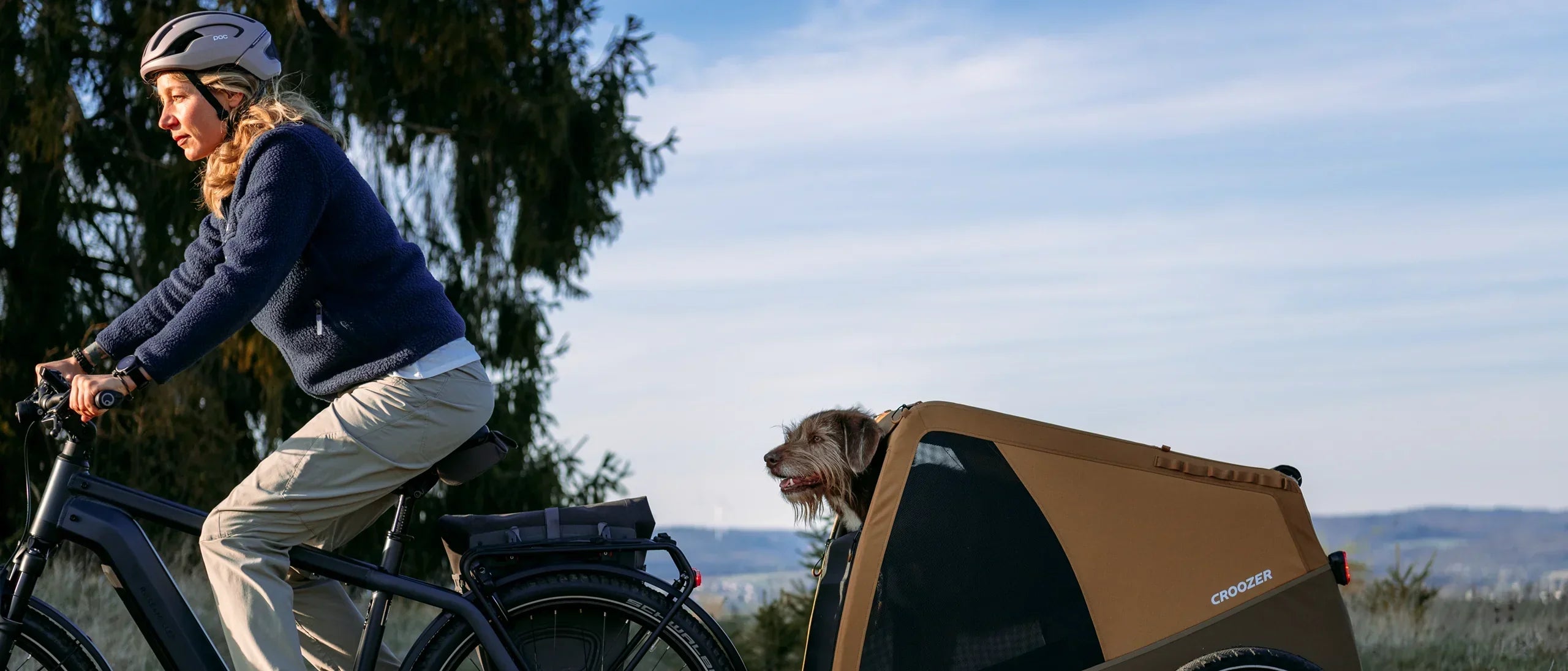 Woman rides bike with Croozer Dog trailer attached to rear wheel; dog sticks head out, landscape behind.