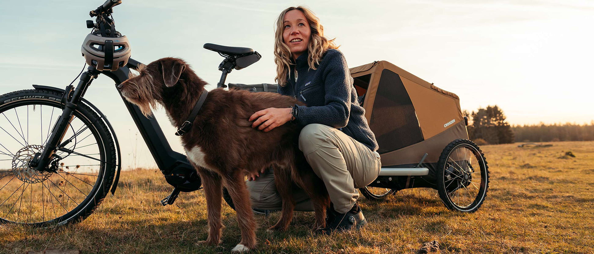 A woman kneels on a grassy meadow, stroking her dog between her legs; a bike with Croozer dog trailer stands nearby; forest and sunset behind.
