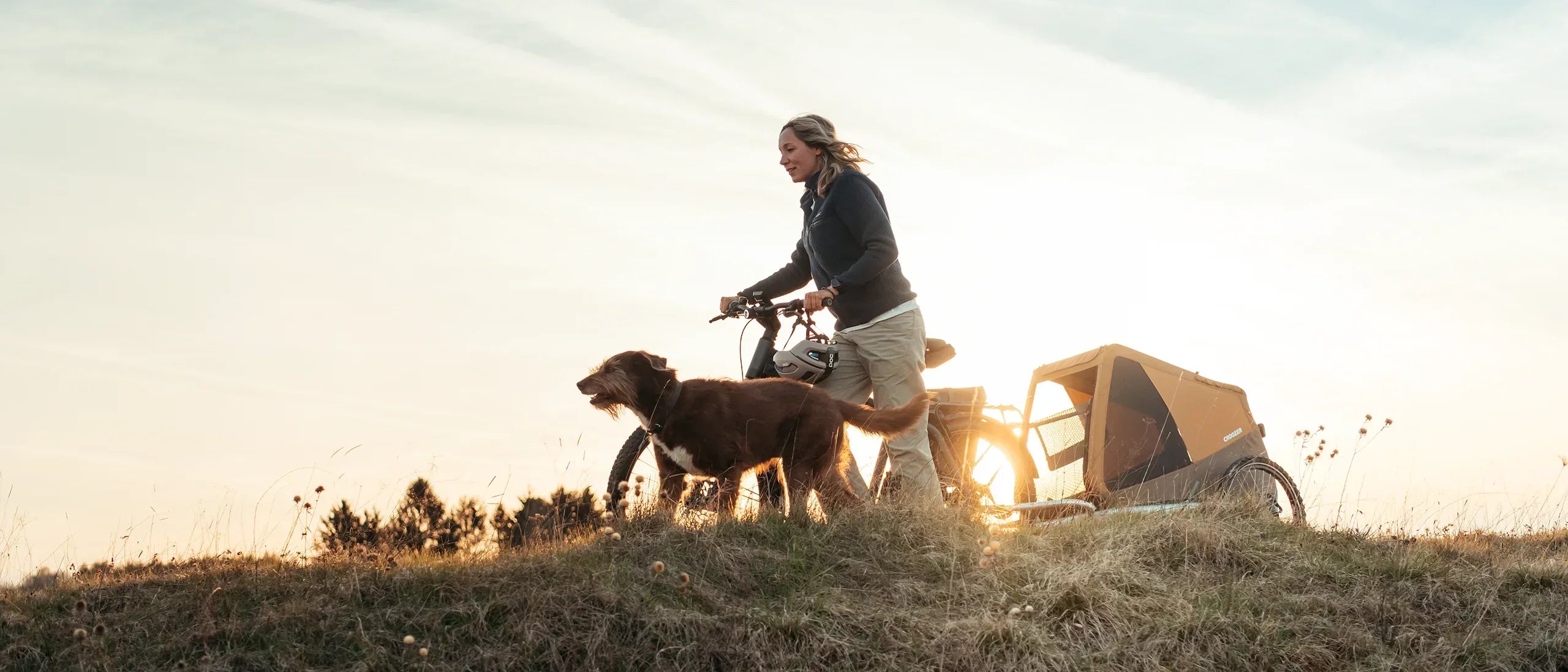A woman and her dog walk across a grassy hill as she holds her bike with a Croozer dog trailer attached; sun setting in the background.