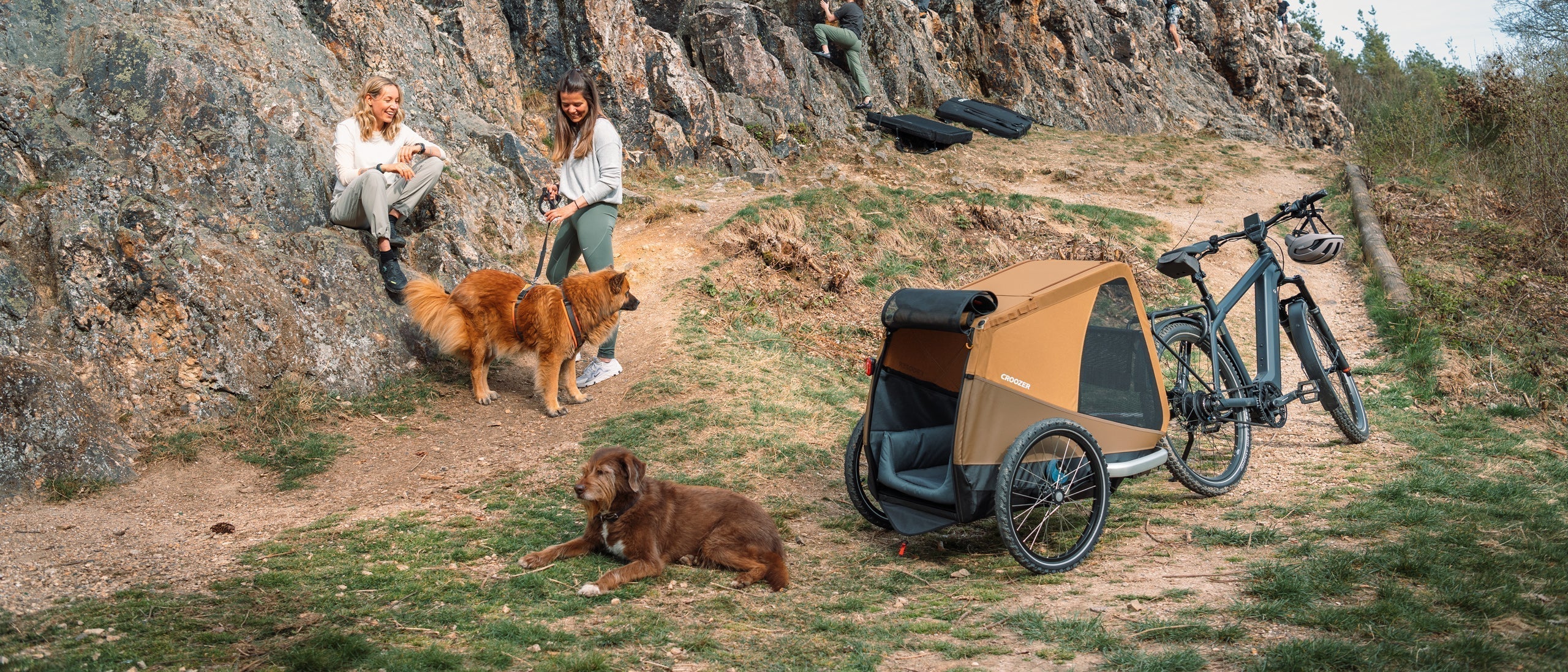Two women with two dogs by a rock; one person sits, the other holds her dog’s leash; another dog rests near Croozer Dog trailer with bed; climbers in background.