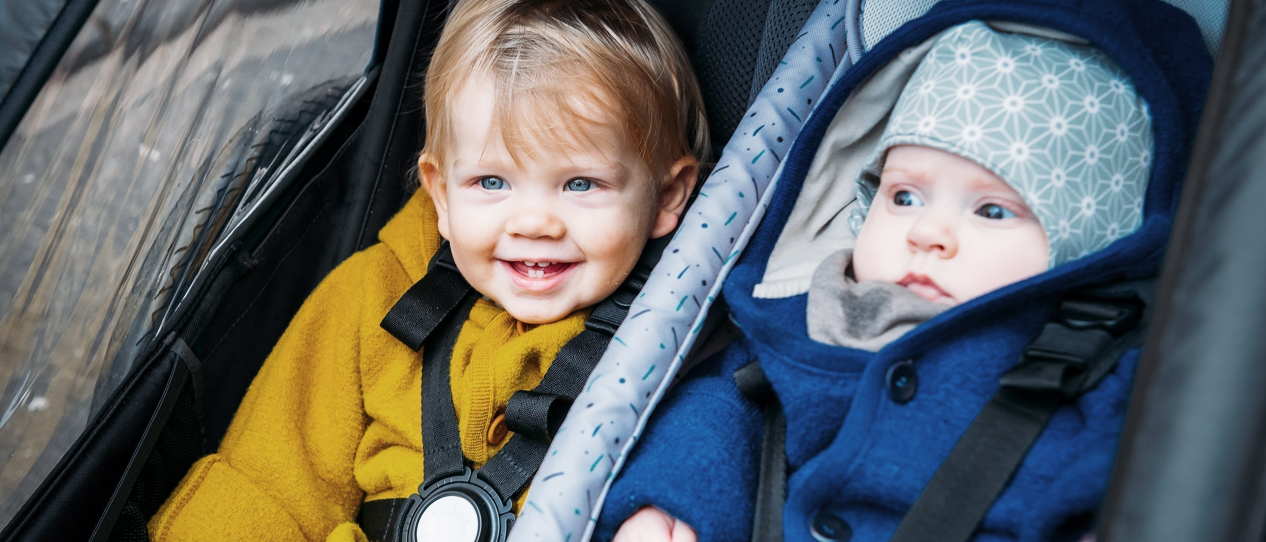 Close-up of two kids in Croozer child trailer: toddler laughing, baby in Croozer baby seat looking sleepy and drowsy.