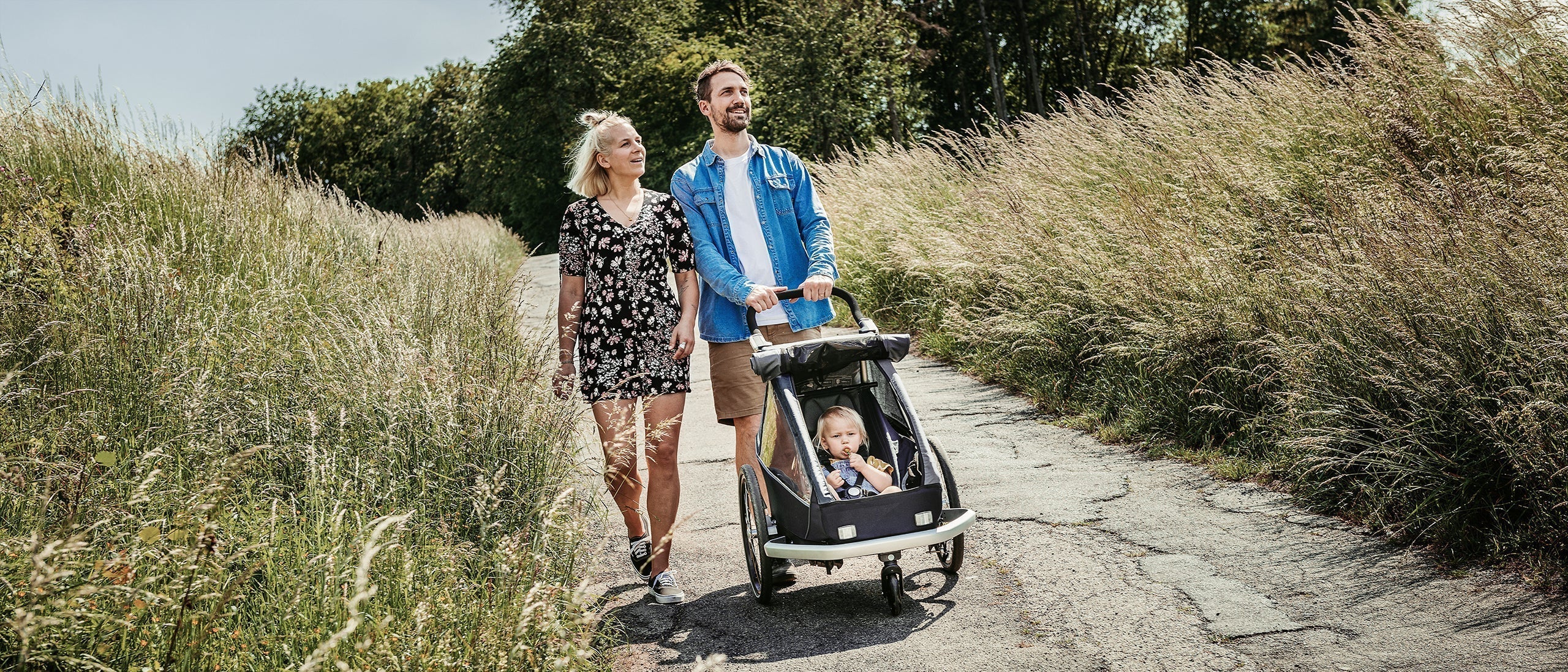 Man and woman push a Croozer Kid Vaaya trailer in buggy mode on a lush path with their child.