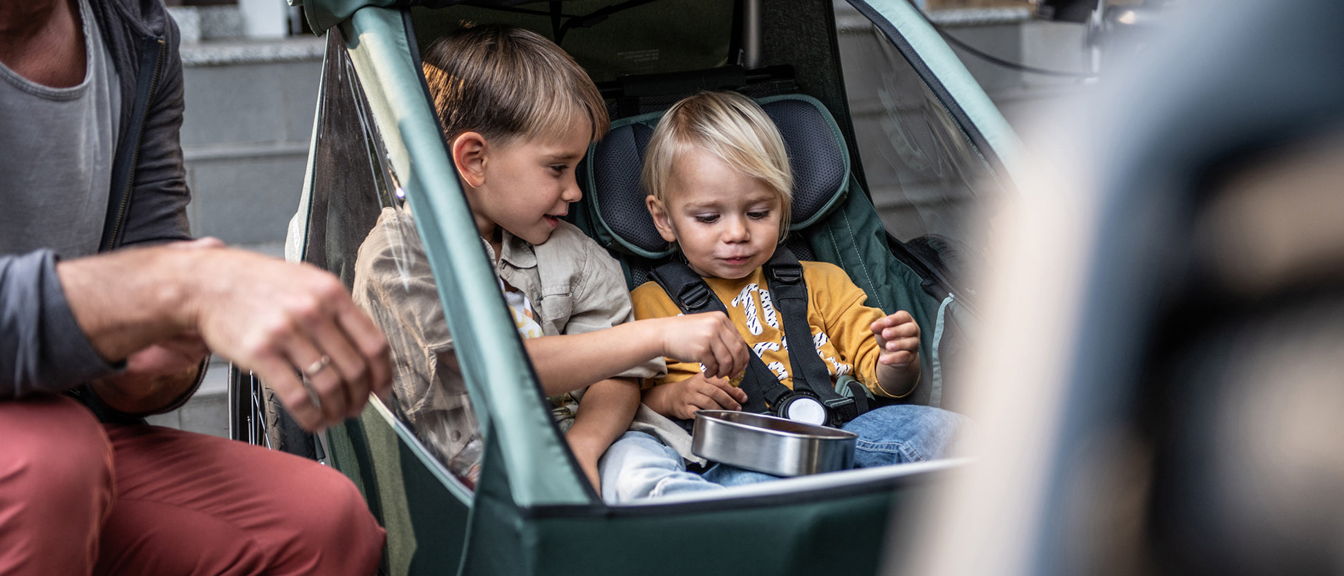 Two children sit in a Croozer child trailer eating gummy bears; dad crouches next to them, joyful atmosphere.