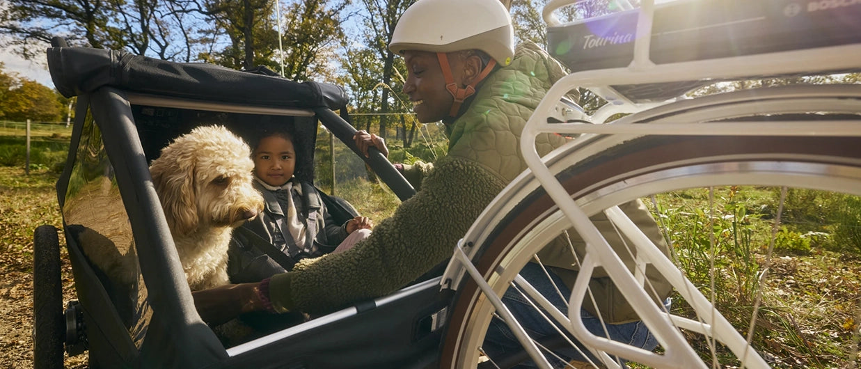 Parent securing dog in Croozer Yuuna while child already secured inside, trailer attached to bike in forest setting