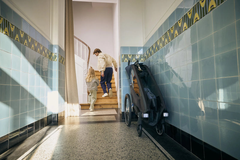 Fahrradanhänger Lykke steht gefaltet im Flur. Im Hintergrund laufen ein Mann und ein Mädchen die Treppe hinauf.