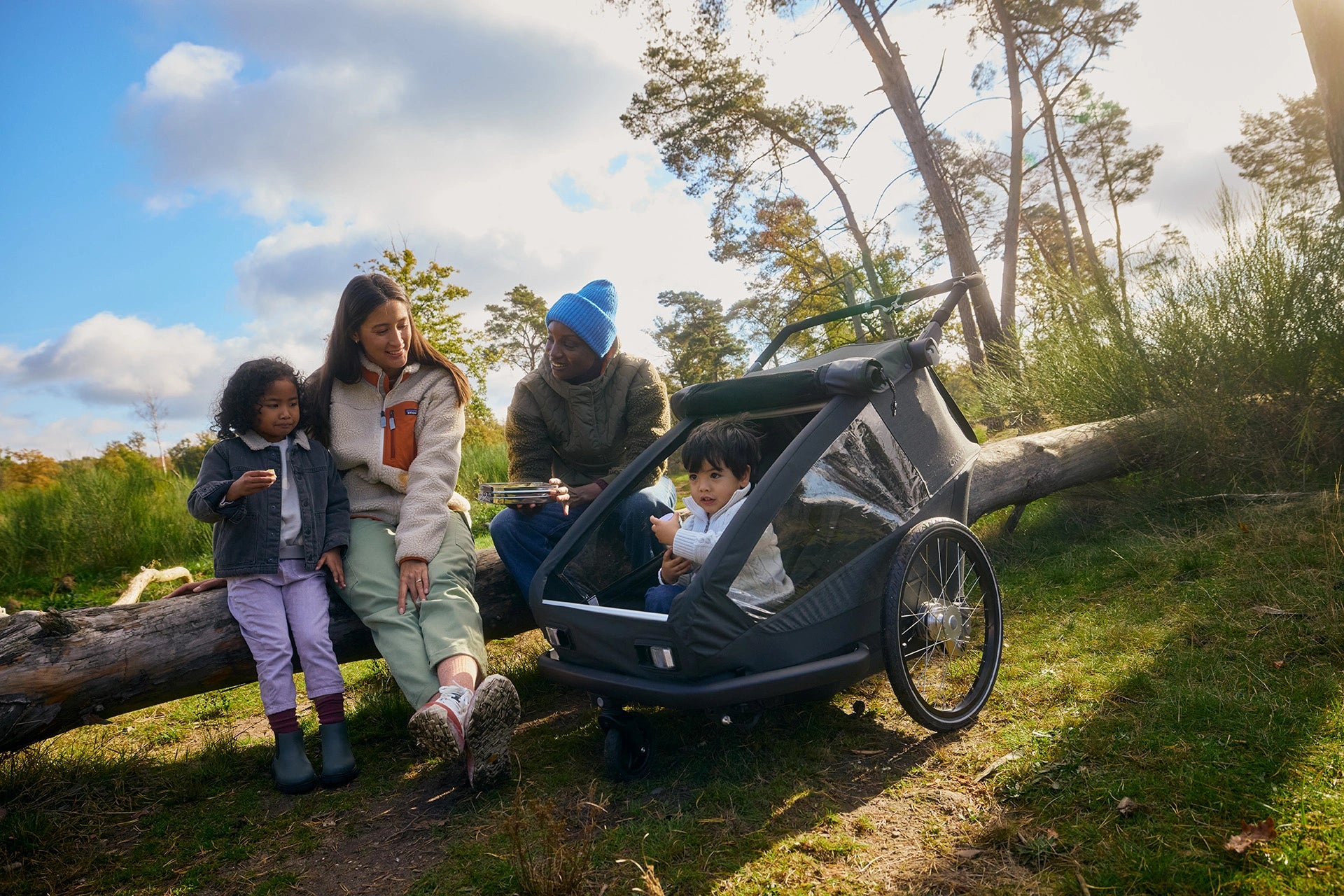 Family enjoying picnic on fallen tree trunk while child sits in Croozer Yuuna trailer, forest environment