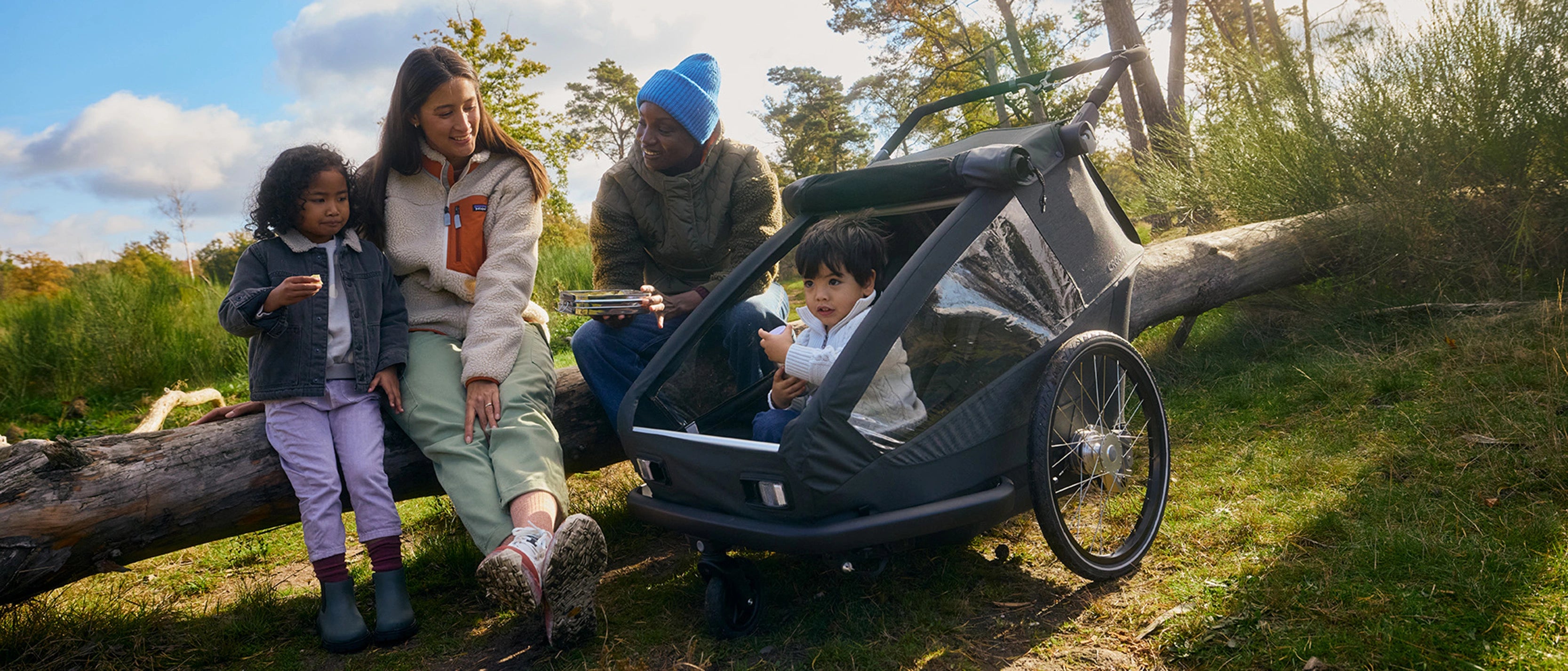 Family enjoying picnic on fallen tree trunk while child sits in Croozer Yuuna trailer, forest environment