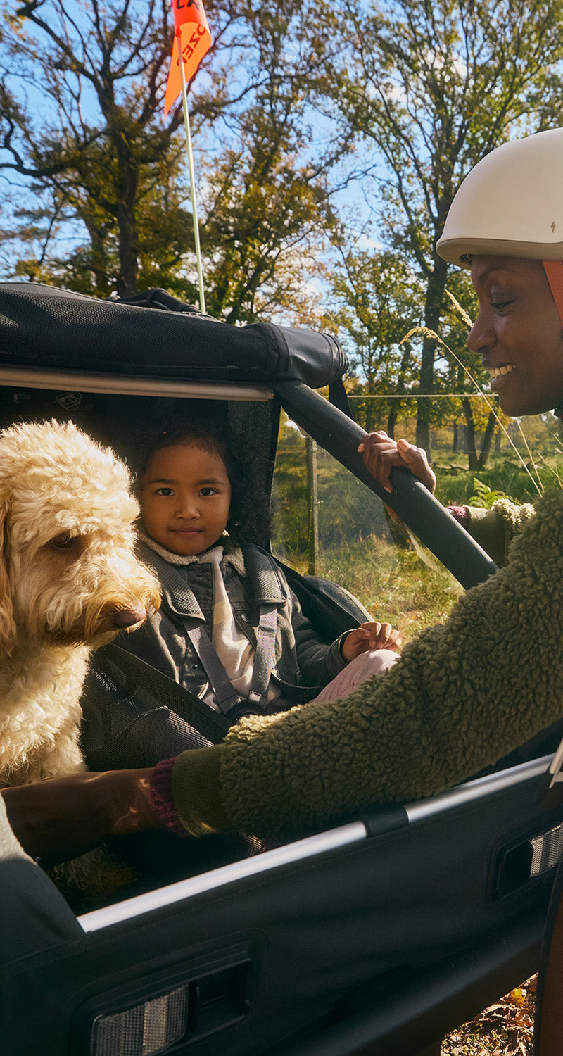 Lächelnde Mutter bei sonnigem Tag unterwegs mit Kind und Hund im Croozer Yuuna