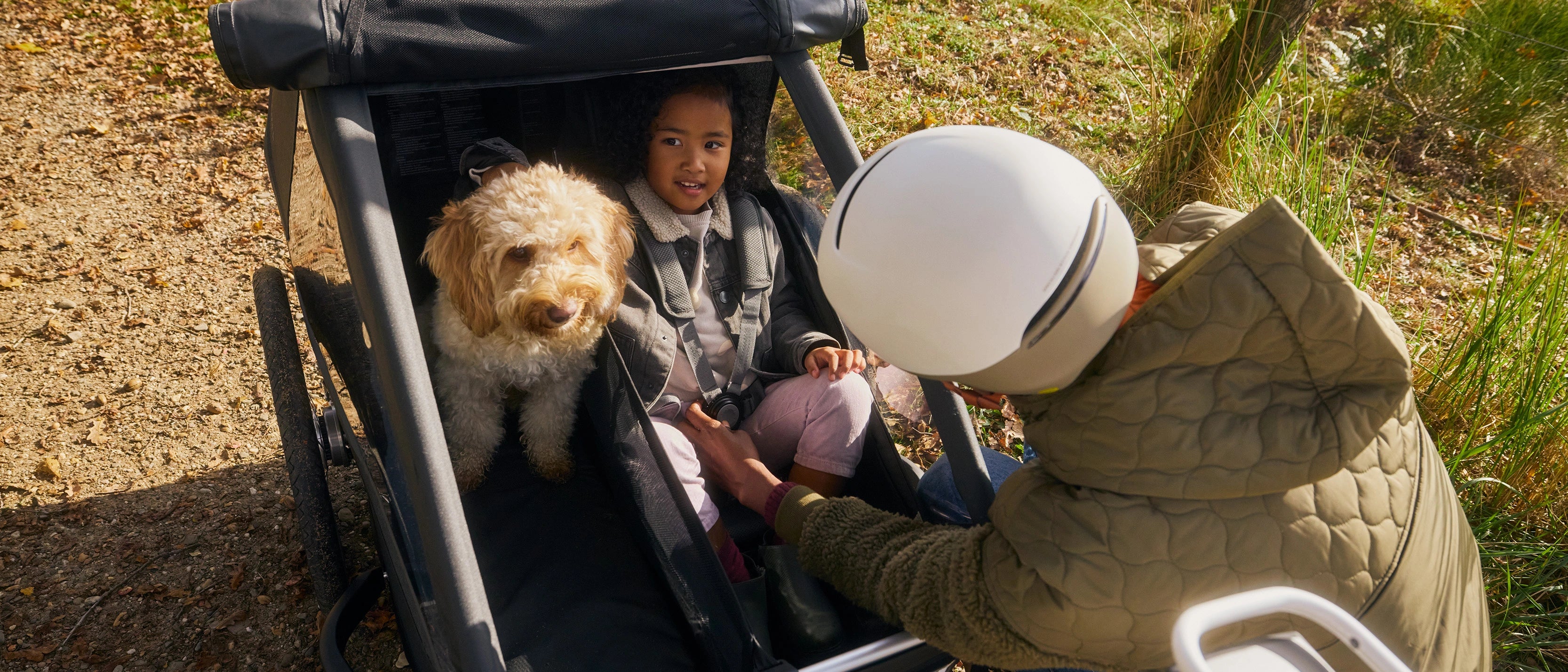Woman with bike helmet securing daughter in Croozer Yuuna modular trailer while dog sits beside her, forest grassland setting