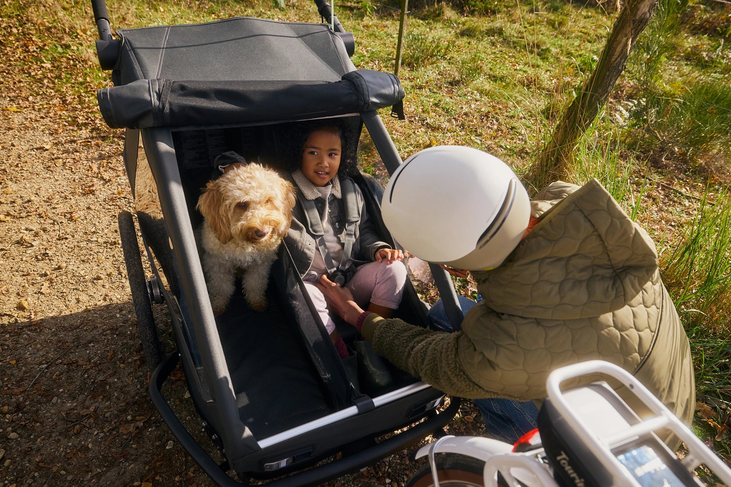 Woman with bike helmet securing daughter in Croozer Yuuna modular trailer while dog sits beside her, forest grassland setting