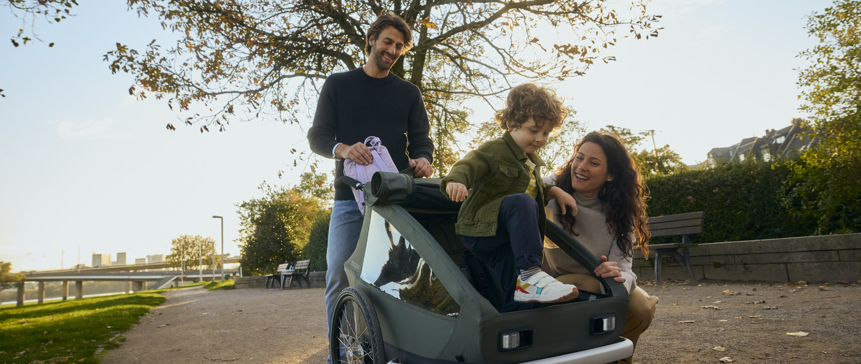 Aus dem Croozer Ellma Kinderanhänger steigt ein Kind aus, die Eltern sind hinter und neben dem Anhänger. 
