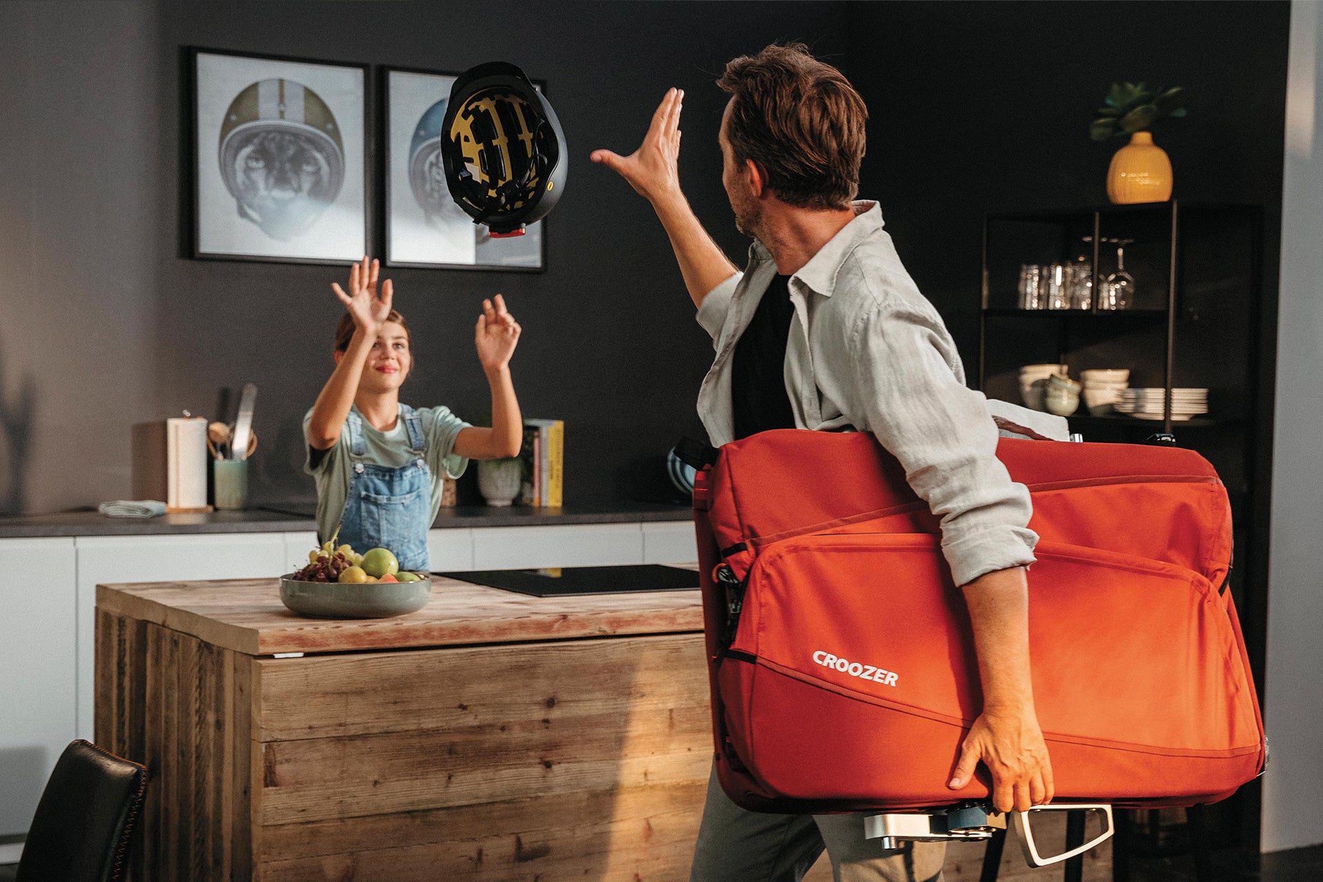 Father and daughter preparing for bike tour with Croozer Cargo bike trailer, passing helmet in kitchen