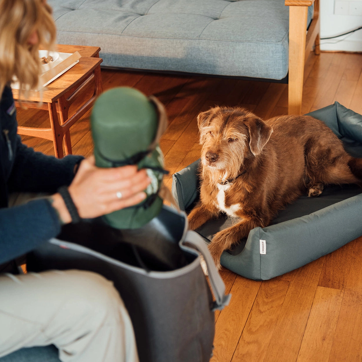 Woman packing bag for outdoor adventure while dog watches from Croozer Dog bed, both in living room