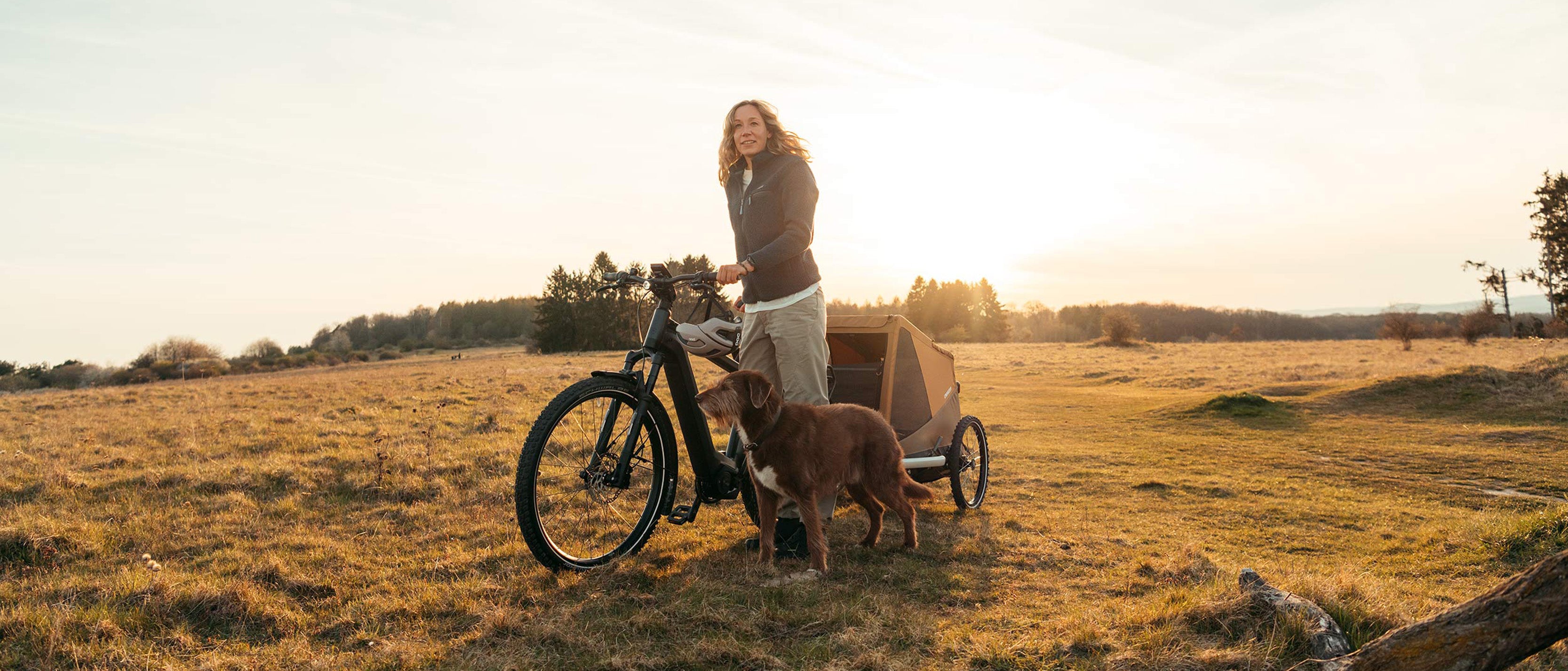 Woman walking with bike while dog walks alongside, Croozer Dog trailer attached - at sunset