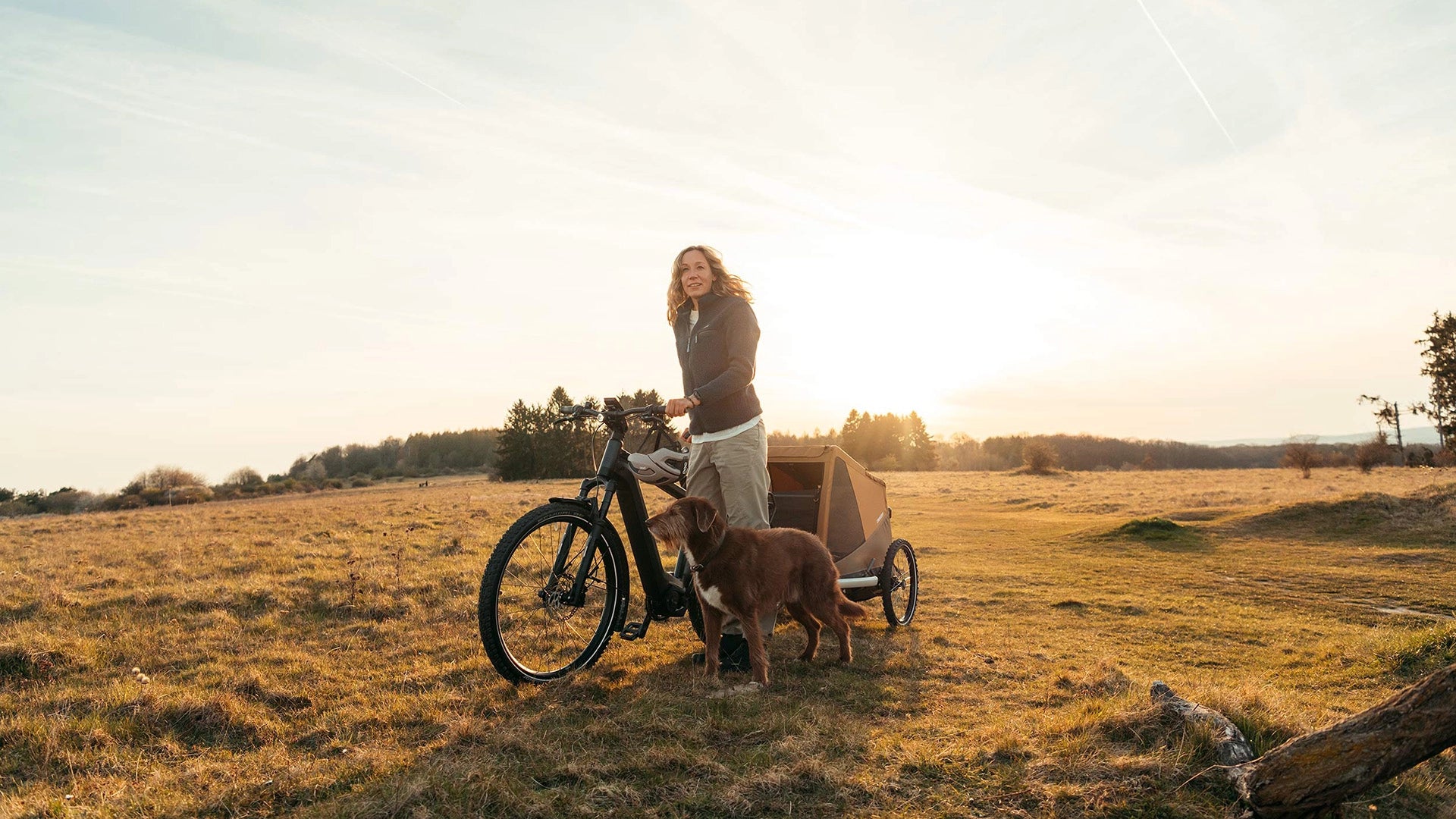 Woman walking with bike while dog walks alongside, Croozer Dog trailer attached - at sunset