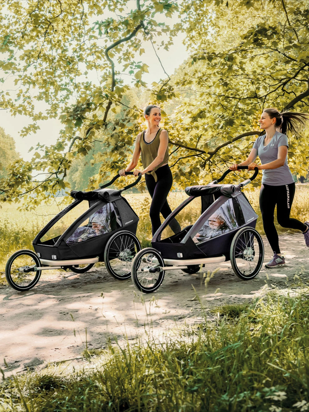 Two women jogging with Croozer Kid Vaaya and Keeke child bike trailers in park setting