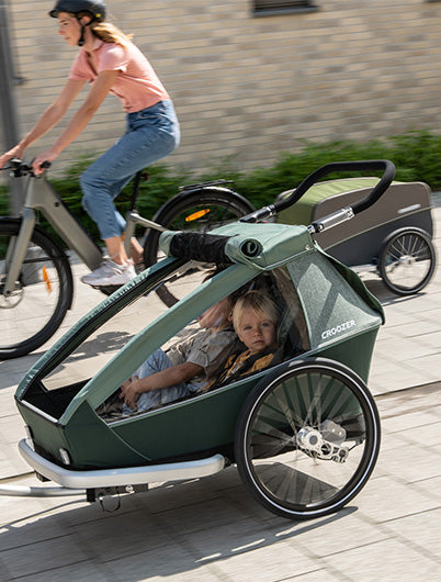 Croozer kid bike trailer with child passenger in foreground woman cycling with Croozer cargo trailer in background