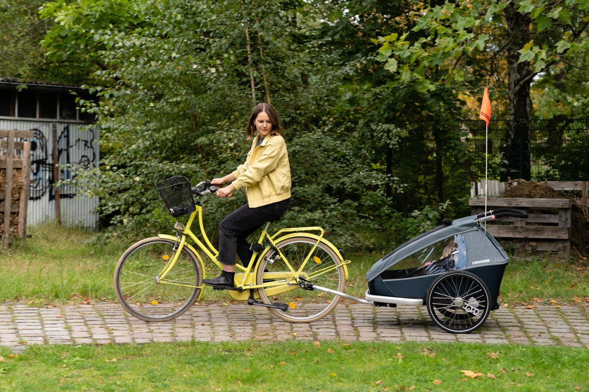Woman in yellow jacket riding yellow bike with attached Croozer Kid Vaaya bike trailer carrying child on cobblestone path