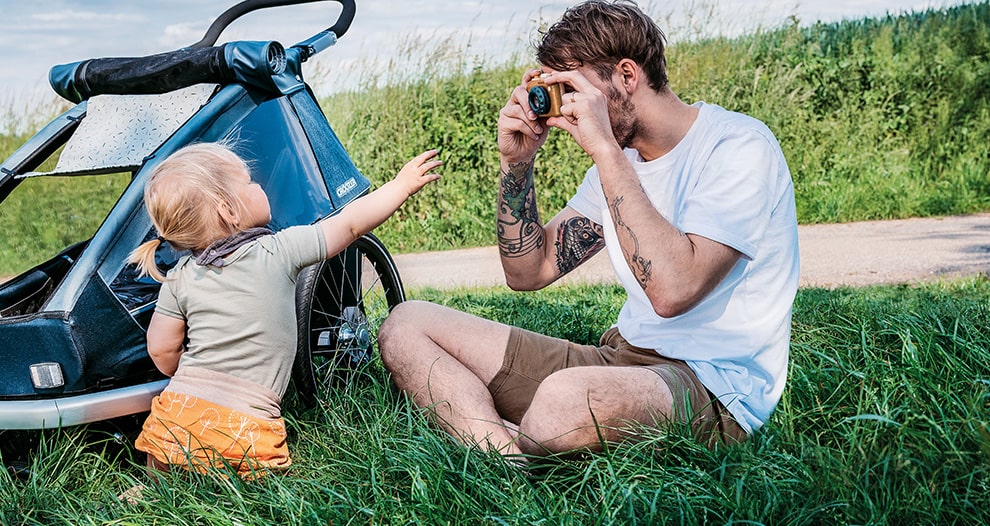 Father and young child sitting in grass next to blue Croozer kid trailer taking photos together in countryside