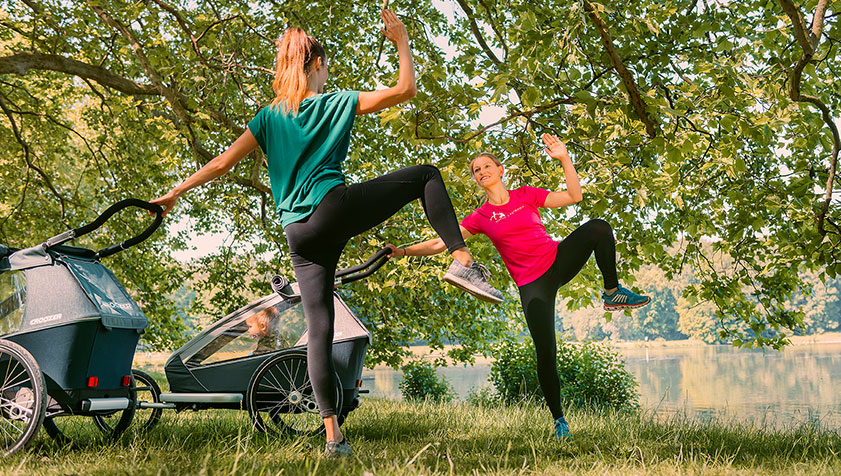Two women doing outdoor fitness exercises with Croozer bike trailer in park setting under trees