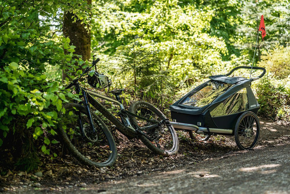 E-bike connected to graphite blue Croozer Vaaya child bike trailer on woodland trail with lush green forest background