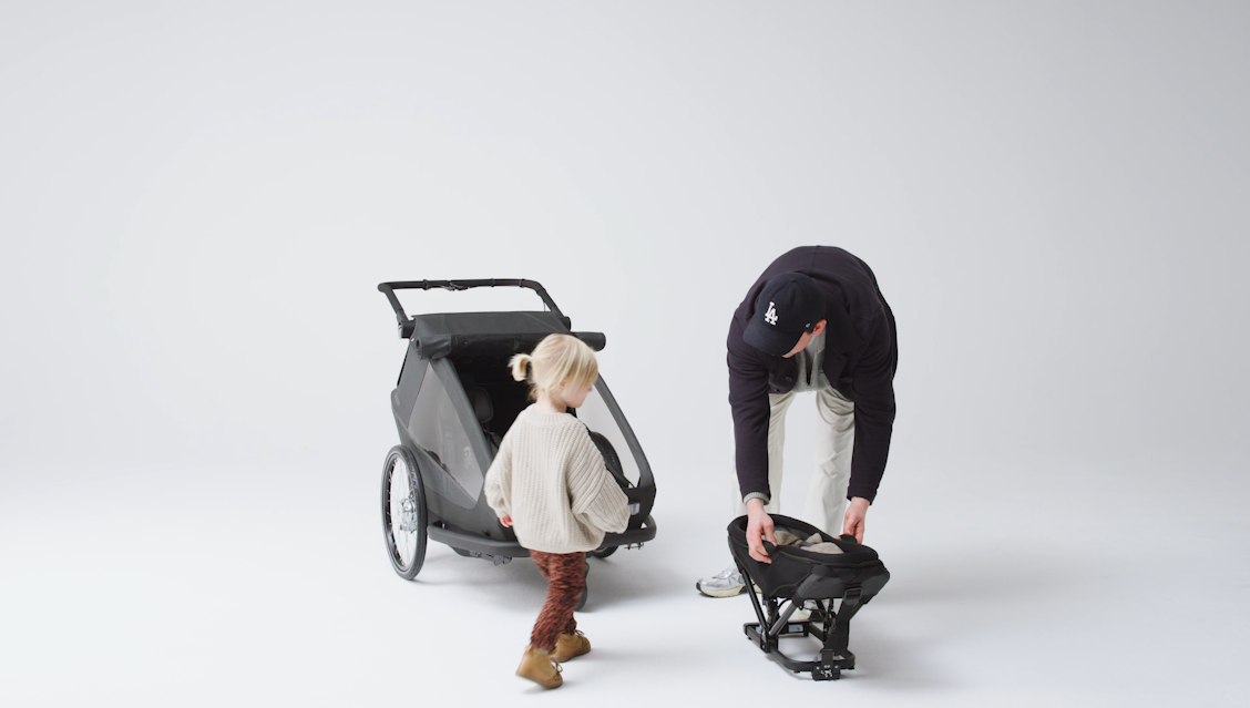 Parent installing baby seat module into Croozer Yuuna modular bike trailer while child watches in studio setting