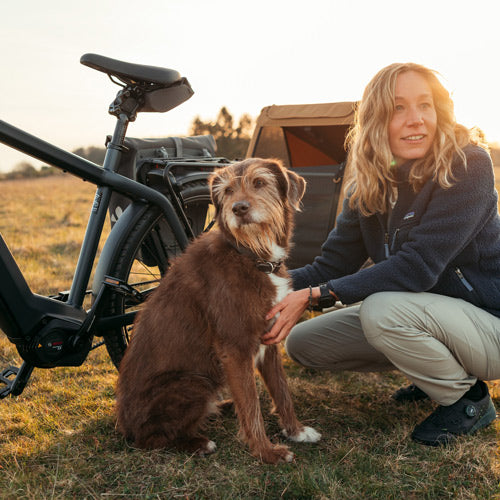 Blonde woman petting brown dog next to electric bike with attached Croozer dog trailer during sunset